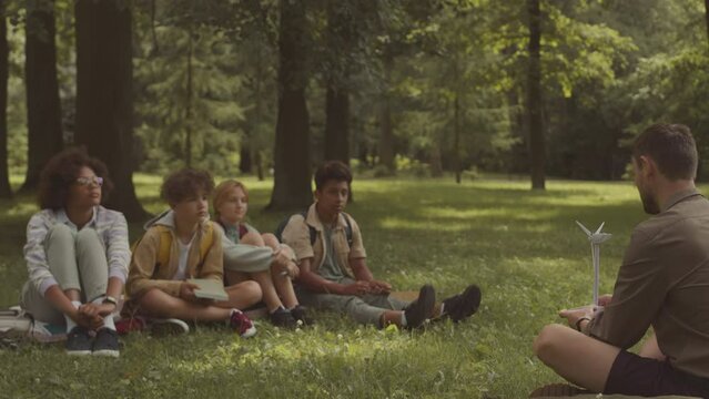Young male teacher with windmill model in hands telling of alternative sources of energy to diverse group of school children sitting on green grass in public park having outdoor class