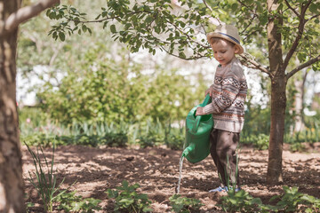 A cute boy in a hat plants and waters seedlings from a watering can in a summer garden, outdoors. The concept of gardening and teaching a child to work.