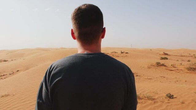 Young man stands in desert in wind and admires dunes. Back view. Concept of solitude and reflection. Slow motion