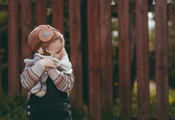 A little boy of 4 years old in an aviator hat and stylish clothes plays outdoors in spring. Portrait of a child with a soft toy in his hands. Happy childhood.