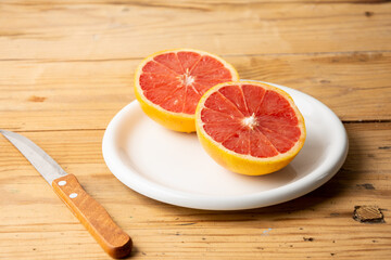Overhead view of split red grapefruit on white plate on wooden table with knife, horizontal, with copy space