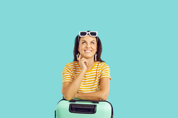 Happy female traveler in anticipation of long-awaited vacation, isolated on light blue background. Joyful dreamy woman in summer clothes looks at copy space leaning on turquoise suitcase. Banner.