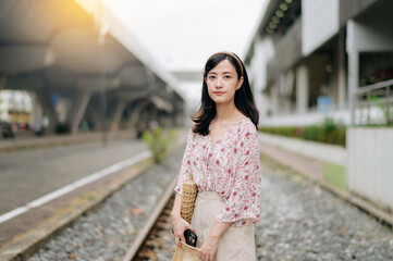 Fototapeta premium young asian woman traveler with weaving basket happy smiling looking to a camera beside train railway. Journey trip lifestyle, world travel explorer or Asia summer tourism concept.