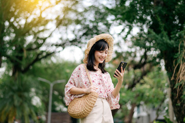 Portrait of asian young woman traveler with weaving hat and basket using mobile phone on green park nature background. Journey trip lifestyle, world travel explorer or Asia summer tourism concept.