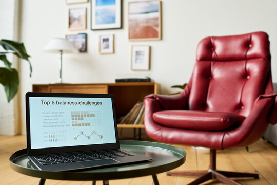Laptop with financial graphic data on screen standing on small round table in front of camera against armchair and pictures hanging on wall
