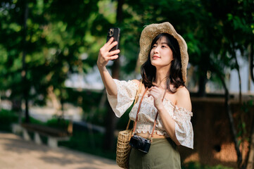 Portrait of asian young woman traveler with weaving hat, basket, mobile phone and camera on green public park background. Journey trip lifestyle, world travel explorer or Asia summer tourism concept.