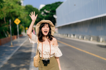 Portrait of asian young woman traveler with weaving hat and basket and a camera waving hand to friend by the street. Journey trip lifestyle, world travel explorer or Asia summer tourism concept.