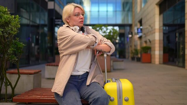 a young female traveler of informal appearance piercing with a suitcase is sitting waiting, looking at a smartphone,being late