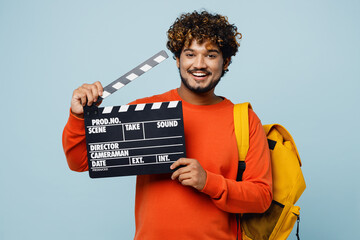 Young teen Indian boy student wear casual clothes backpack bag hold classic black film making clapperboard isolated on plain pastel light blue cyan background. High school university college concept.