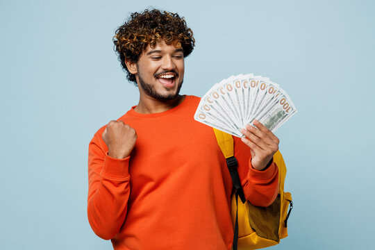 Young Teen Indian Boy Student Wears Casual Clothes Backpack Bag Hold Fan Of Cash Money In Dollar Banknotes, Do Winner Gesture Isolated On Plain Blue Background. High School University College Concept.