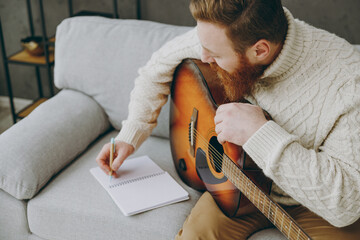 Top view young ginger man wear casual clothes play guitar writing lyrics in notebook sits on grey...