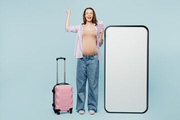 Full body young pregnant mom woman with belly tummy wear casual clothes hold bag passport ticket big huge blank screen area mobile cell phone isolated on plain blue background. Tourist travel abroad