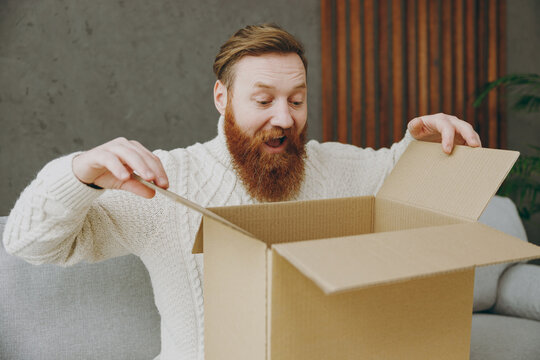 Young surprised shocked happy ginger man wear casual clothes unpacking cardbox box sit on grey sofa couch stay at home hotel flat rest relax spend free spare time in living room indoor Lounge concept