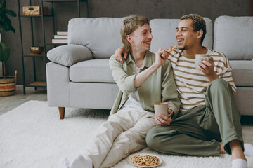 Young fun couple two gay men wear casual clothes together eat cookies feed boyfriend sit on sofa...