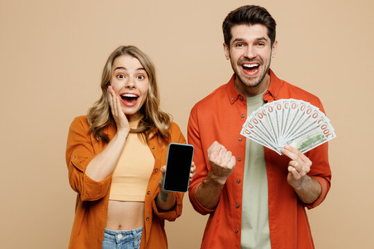 Young couple two friends family man woman wear casual clothes hold in hand fan of cash money in dollar banknotes use mobile cell phone together isolated on pastel plain light beige background studio.