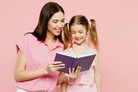 Happy smiling woman wear casual clothes with child kid girl 6-7 years old. Mother daughter read book together before going to bed isolated on plain pastel pink background. Family parent day concept.