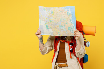 Young woman carrying backpack with stuff mat covering face with map isolated on plain yellow...