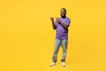 Full body young man of African American ethnicity wear casual clothes purple t-shirt point index finger aside indicate on workspace area copy space mock up isolated on plain yellow background studio.