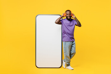 Full body young man of African American ethnicity wears casual clothes purple t-shirt big huge blank screen mobile cell phone with area liste music in headphones isolated on plain yellow background.