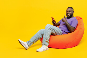 Full body fun young man of African American ethnicity wear casual clothes purple t-shirt sit in bag chair hold in hand use mobile cell phone show thumb up isolated on plain yellow background studio.