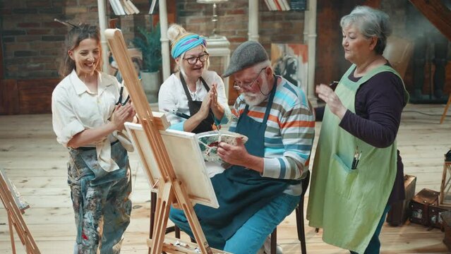 Colleagues and artist friends applaud and support an elderly man who successfully completes his work by painting on a canvas in a group art class 
