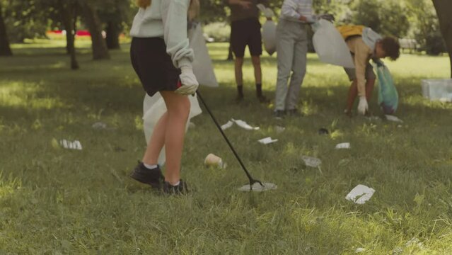 Caucasian schoolgirl with multiethnic classmates and male teacher collecting rubbish on green grass in summer park