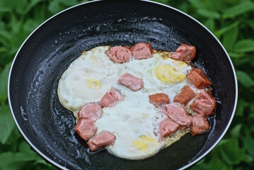 a meal of fried eggs with pieces of red sausages in a black pan on a green background