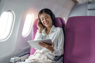 Young asian woman in wireless headphones listening to music and smiling during fly in airplane,...