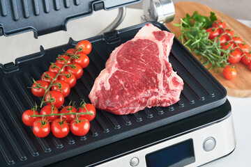 Raw prime steaks on electric grill. Black angus prime meat steaks Ribeye on modern electric grill with rosemary, cherry tomato and pepper. Cooking process in modern kitchen. Top view, copy space.