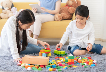 Fototapeta premium wo kids sitting on living room floor play with colourful block toy construction game. Asian little girl enjoy creative activity building development with her sibling at home. Family leisure together.