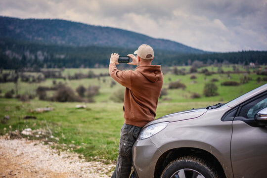 Young adult male traveler standing next to car making landscape photo using smartphone. Travel to mountains