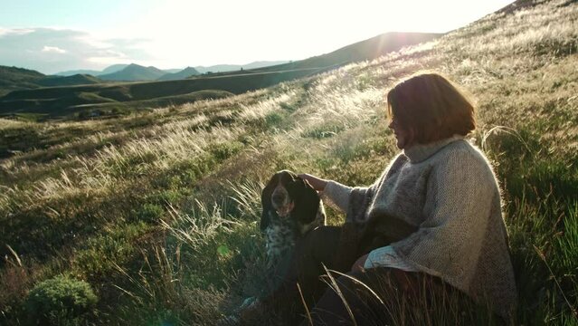 A woman caresses her dog against the backdrop of a mountain range in the rays of the sunset. The feather grass shines. Charming landscape