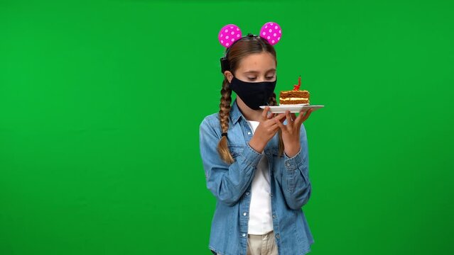 Teen Caucasian girl in coronavirus face mask with piece of birthday cake gesturing helplessness looking at camera. Sad teenager posing on green screen celebrating holiday on Covid-19 pandemic
