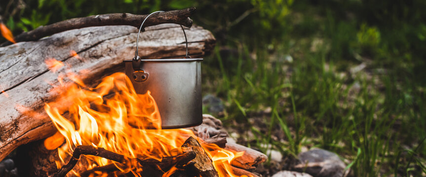Kettle hanging over fire. Cooking food at fire in wild. Beautiful big log burns in bonfire close-up. Survival in wild nature. Wonderful flame with caldron. Pot hangs in flames. Campfire background.