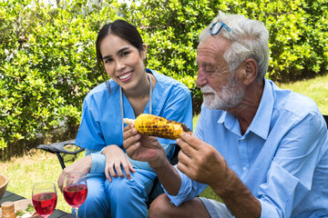 Elderly man eating grill corn with smiling care giver woman sitting besides in Senior Home Care