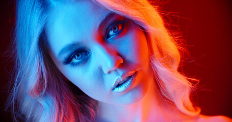 Sexy caucasian blonde girl look at camera with sensual stare, weariing bright glowing makeup, standing in red and blue neon lights - nightlife, cyberpunk footage close up portrait
