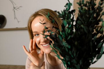 Portrait of a Cute young blonde woman in a beige top behind a plant leaves, happy smiling, Japanese style studio at the background