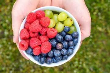 pełna miska owoców trzymana w dłoni na tle zielonej trawy, borówki winogrona maliny 