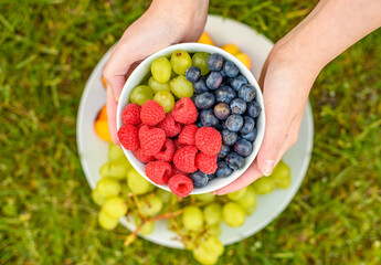 Miseczka z borówkami winogronem i malinami trzymana w rekach na tle talerza z owocami na zielonym...