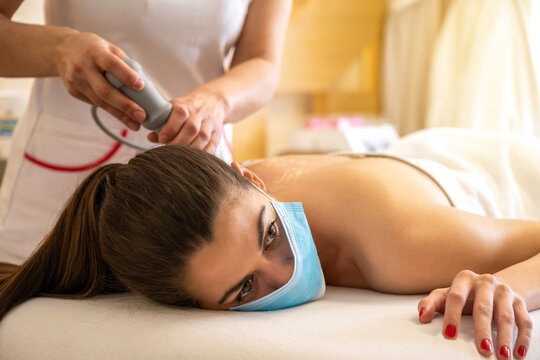Young woman wearing protective mask having extracorporeal shockwave therapy in beauty salon.