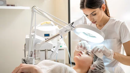 Keuken foto achterwand Schoonheidssalon Beautician squeezing acne and pimples from young woman's nose using lamp and magnifying glass in beauty spa clinic.