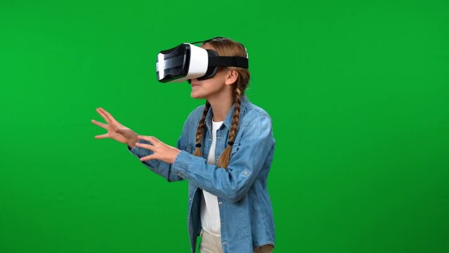 Excited teen Caucasian girl gaming online in VR goggles turning at green screen background. Absorbed happy teenage gamer enjoying video game at chroma key