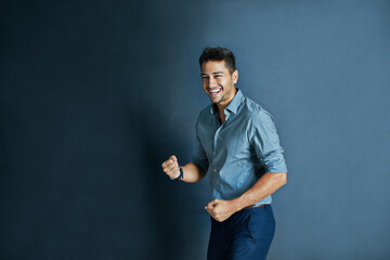 Celebration, fist pump and portrait of a businessman in a studio with achievement or winning. Happy, smile and professional male employee winner with success to celebrate isolated by blue background.