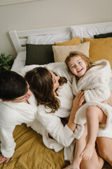 Young mother, father hugs cute little daughter in bathrobes lying on bed, look at each other in hotel room. Happy mom, dad and girl kid face to face and smiling. Top view. Family with child having fun