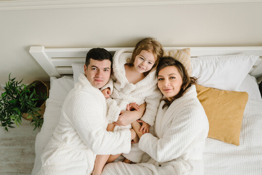 Young mother, father hugs cute little daughter in bathrobes lying on bed, look at camera in hotel room. Healthcare concept. Happy mom, dad and girl kid smiling top view. Family with child having fun.