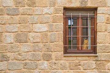 Fototapeta na wymiar The wall of the old house with a window, the cat looks out the window, the streets of the medieval city of Rhodes, beginning of the season of travel to popular places in Dodecanese archipelago Greece