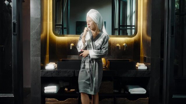 Cleaning teeth. Young woman with towel on her head stand in modern bathroom with big mirror. Big house with dark interior. Rich owner of luxury apartment, real estate concept