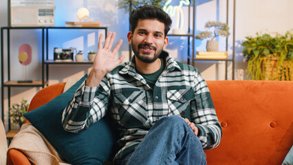 Hello. Indian handsome man smiling friendly at camera and waving hands gesturing hello, hi, greeting or goodbye, welcoming with hospitable expression at home. Hindu guy sitting on sofa in living room