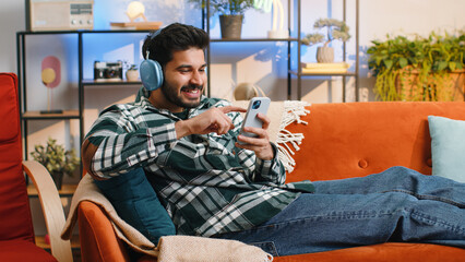 Happy overjoyed hindu indian man in wireless headphones relaxing sits on couch at home apartment choosing listening favorite energetic disco dancing music. People weekend daytime leisure activities
