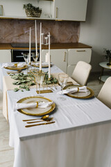 Festive table setting with plate, golden cutlery set and eucalyptus leaves, candles. Dinner table decorated for cozy fall holiday gathering in kitchen. Modern panel kitchen interior with countertop.